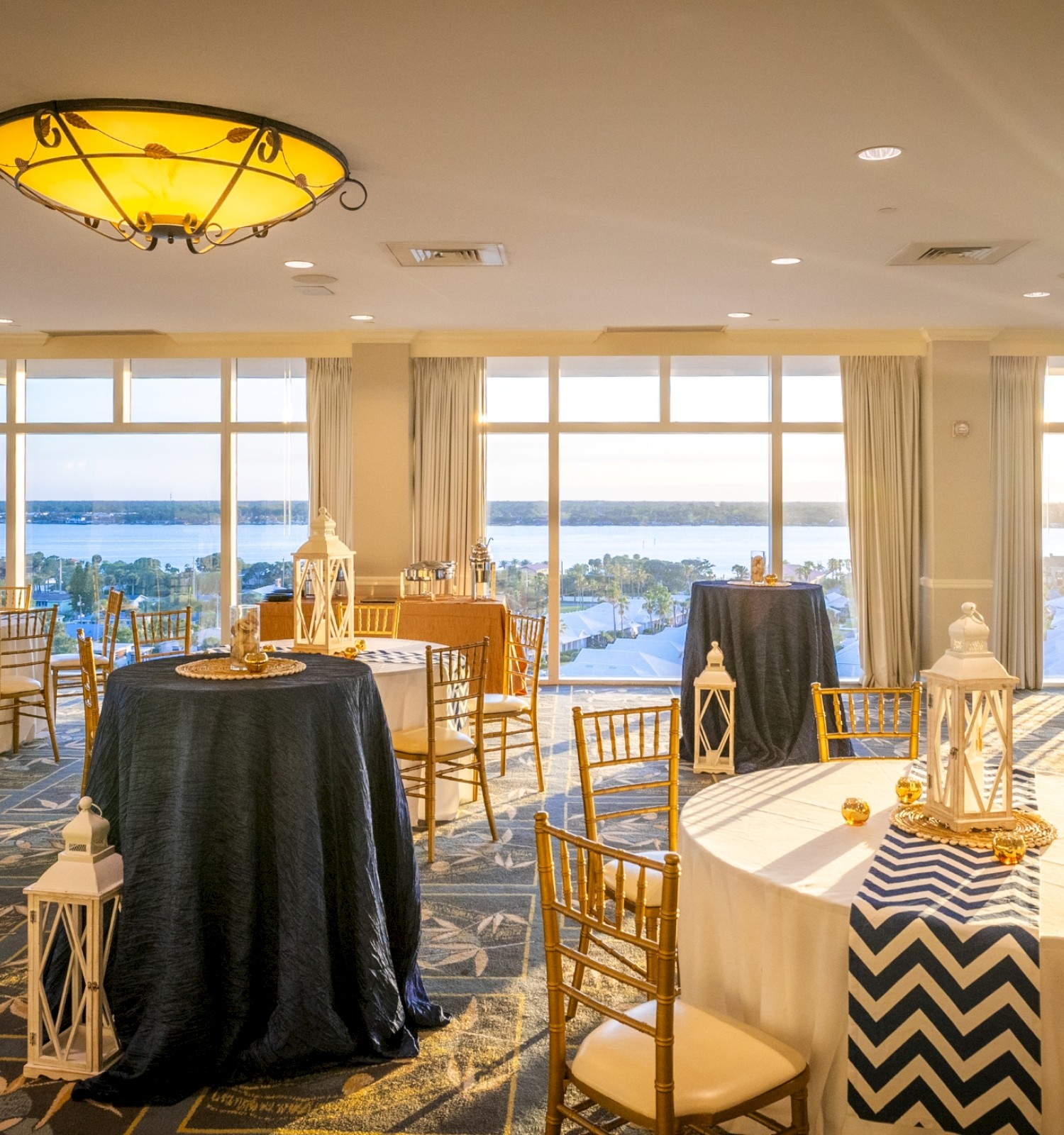 A bright event space with large windows, round tables draped in black and white, gold chairs, lantern centerpieces, and warm sunset light.