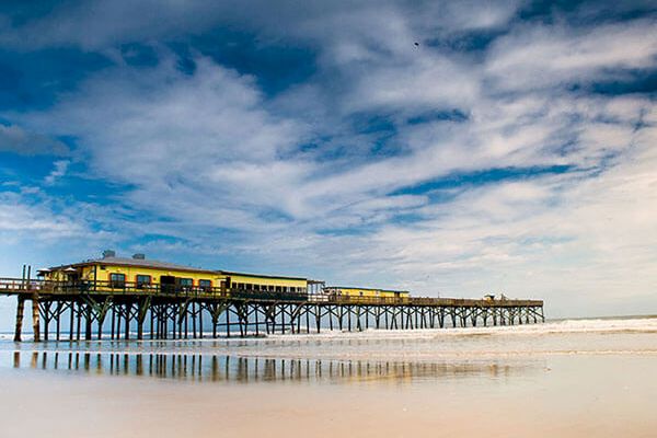 The image shows a long pier extending over a calm beach with a vibrant blue sky and scattered clouds in the background.