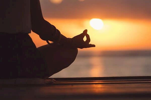 A person is sitting in a meditative pose by the water during sunset, with the focus on their hand in a mudra gesture and the serene background.