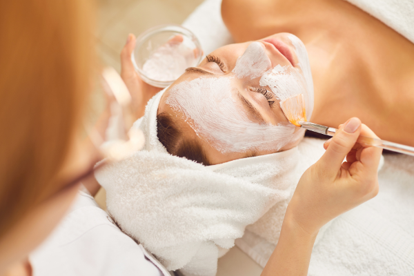 A person is receiving a facial treatment, with a mask being applied to their face using a brush. They are lying down, covered in a towel.