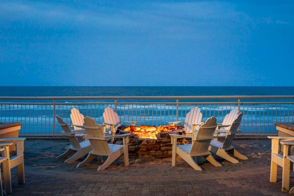 A cozy fire pit with a ring of white Adirondack chairs overlooks a serene ocean view at dusk, creating a relaxed and inviting atmosphere.