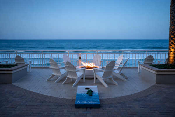 A cozy seaside patio with Adirondack chairs around a fire pit, overlooking the ocean at dusk, creating a peaceful, inviting atmosphere.