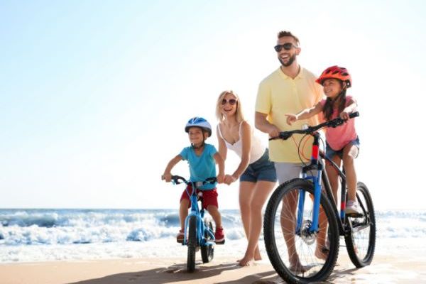 A family is riding bicycles on a sandy beach next to the ocean; the sun is shining, and everyone appears happy and enjoying the day.