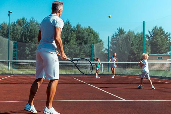 A family is playing tennis on an outdoor court, with a man in the foreground preparing to hit the ball while others watch or participate.
