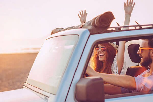 Two people are enjoying a drive in a convertible near the beach, with one person raising their arms in excitement and both wearing sunglasses.