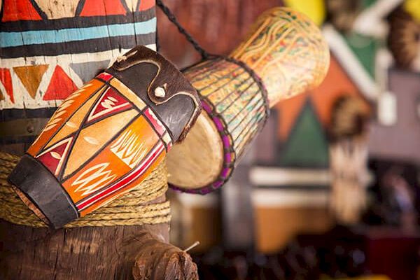 The image shows colorful traditional drums with vibrant patterns hanging on a wooden structure, with a blurred background.