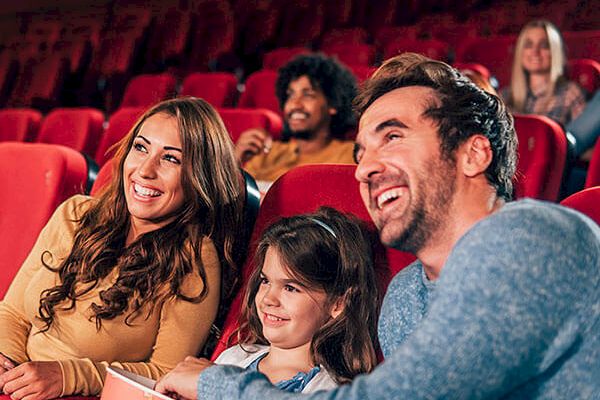 A group of people, including a smiling family with a young girl, are sitting in a movie theater, enjoying a film together.