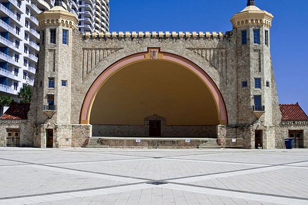 A large outdoor stage with a central archway, flanked by two cylindrical towers, situated in an open plaza with a clear blue sky.