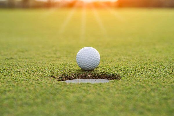 A golf ball is positioned on the edge of a hole on a putting green, with the sun setting in the background.
