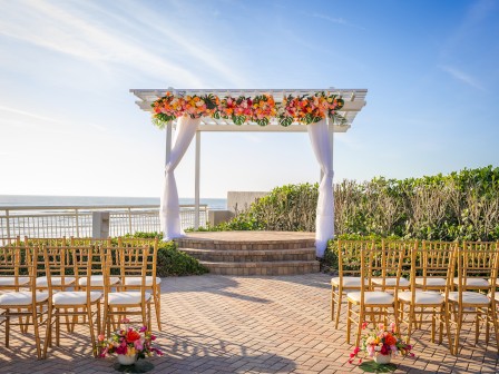 An outdoor wedding setup features a decorated arch, arranged chairs, and floral arrangements with a scenic view, likely by the sea.