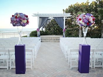 This image shows an outdoor wedding setup by a beach, with white chairs arranged in rows, and two floral arrangements on purple stands flanking the aisle.