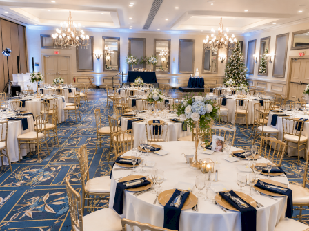 This is an elegant banquet hall decorated for a wedding or formal event with gold chairs, white tables, floral centerpieces, and Christmas trees.