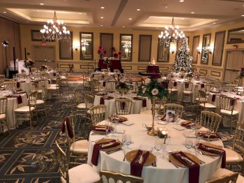 An elegant banquet hall decorated for an event with round tables set with red napkins, gold chairs, and floral centerpieces, under chandeliers.