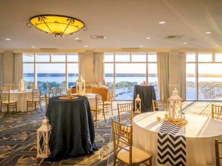 A beautifully decorated event space with round tables, elegant chairs, lanterns, and a view of the water, lit by the setting sun.