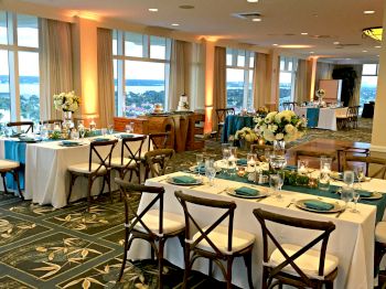 A banquet hall setup with elegant table settings, floral arrangements, panoramic windows, and a view of the water outside, suitable for an event.