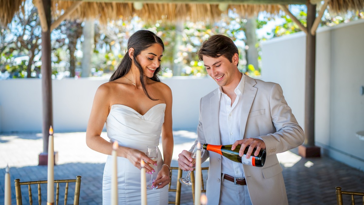 A couple is enjoying an elegant outdoor setting, with the man pouring champagne into a glass. Candles and a festive atmosphere are present.