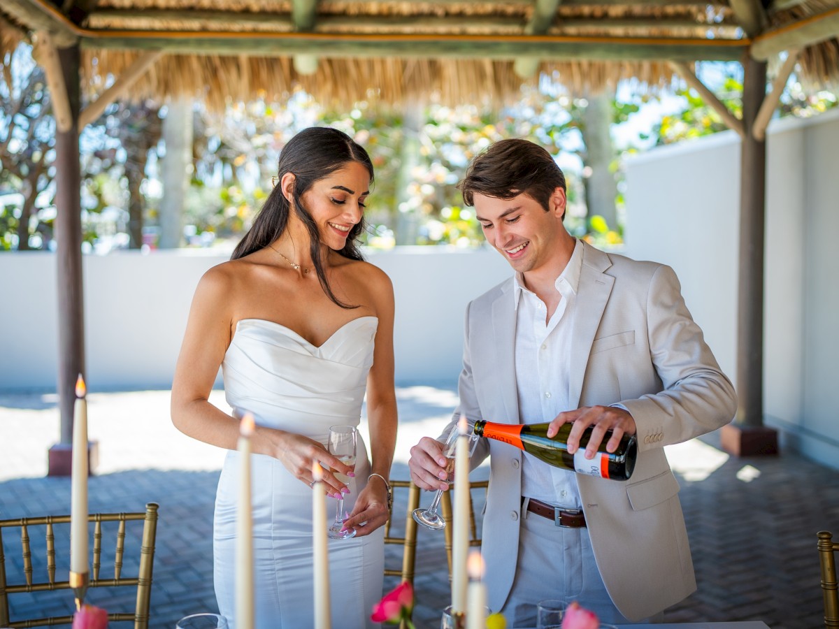 A couple is enjoying an elegant outdoor setting, with the man pouring champagne into a glass. Candles and a festive atmosphere are present.