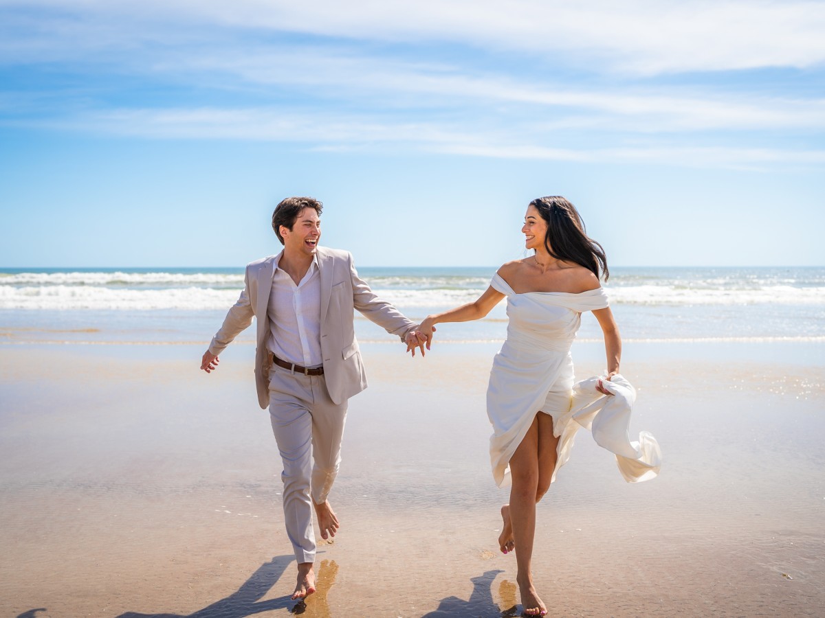 A couple is running barefoot along a beautiful beach, holding hands. The man is in a light suit, and the woman is in a white dress, with the ocean in the background.