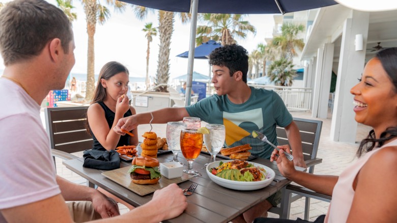 Four friends enjoy a sunny restaurant patio: burgers, salad, drinks; laughter and conversation as they share a meal by the sea.