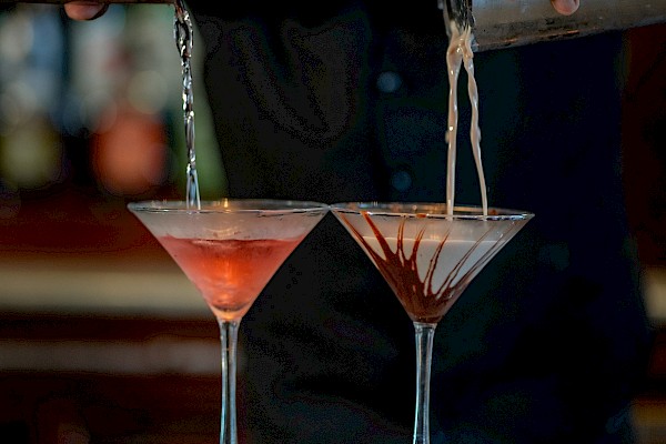 A bartender pours two pink cocktails from a shaker onto a bar, hands poised, glass rims catching the light, ready to serve.