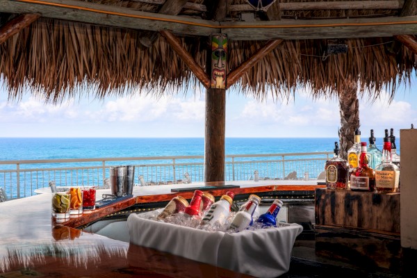 A beachside bar under a thatched roof, with bottles on a curved counter, a chilled tray of drinks, and the ocean view beyond.