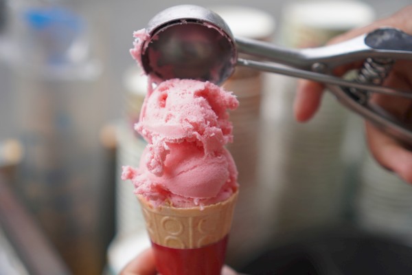 A scoop of pink ice cream being scooped into a cone with a metal scoop, held by a gloved hand.