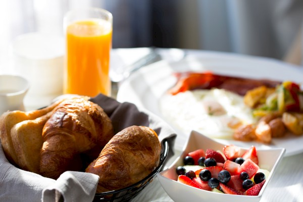 A breakfast spread: croissants in a basket, a glass of orange juice, a plate with eggs and sides, and a bowl of fresh berries.