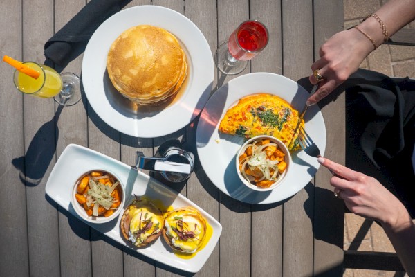 A top-down view of a dining table with brunch: pancakes, omelette, chips, sliders, a glass of rosé, and two people cutting and sharing.