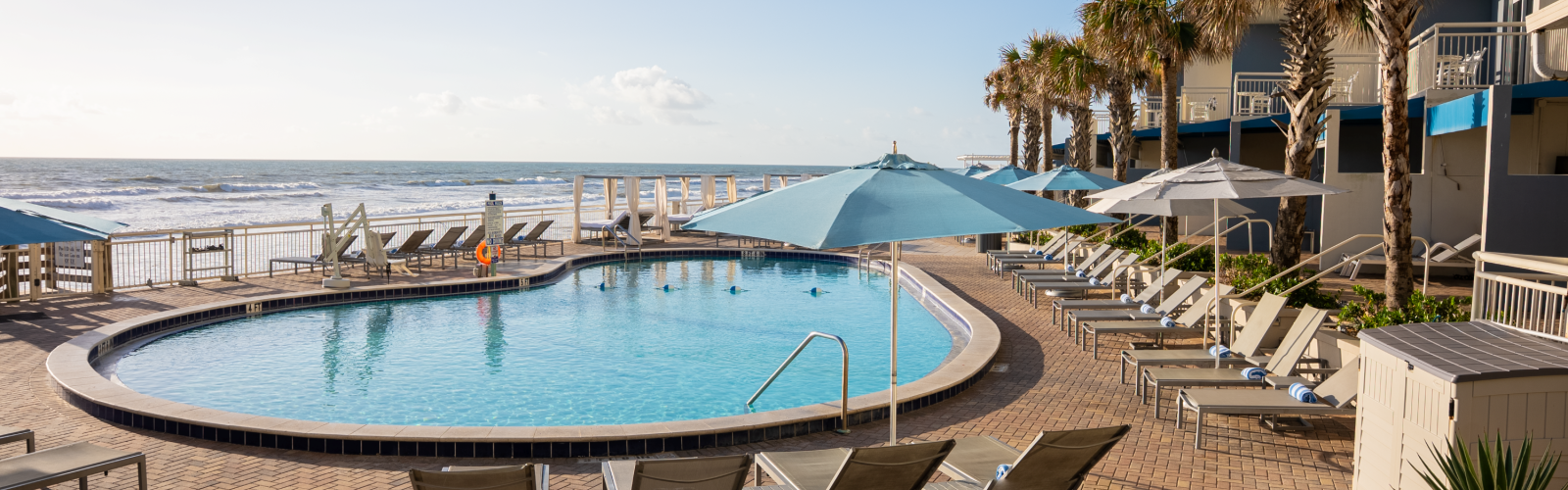 A serene beachfront pool area with chairs, umbrellas, palm trees, and ocean waves in the background on a sunny day.