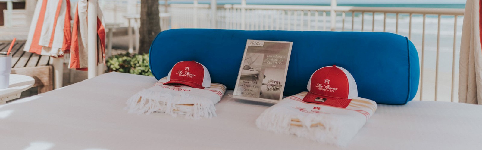 Two red caps, two folded white towels, and a promotional pamphlet are laid out on a bed or lounge chair with a blue cushion, outdoors.