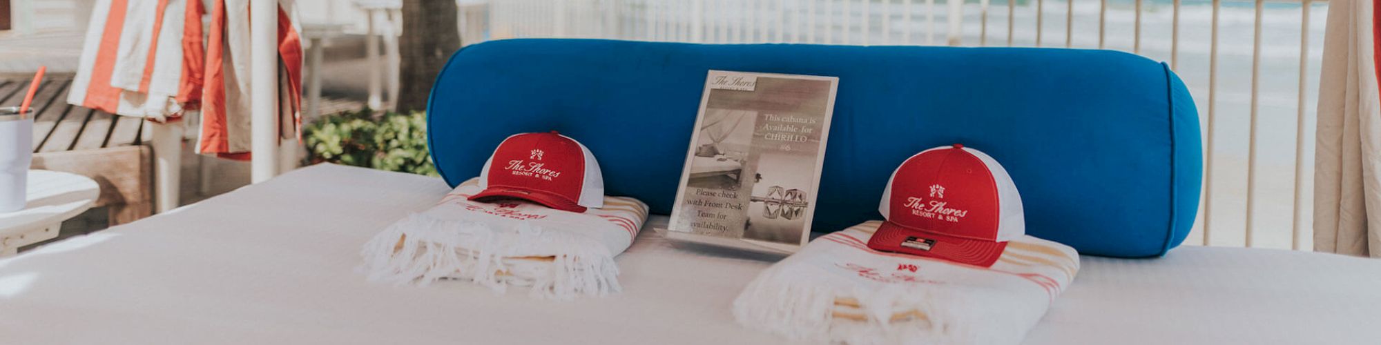 Two red caps, two folded white towels, and a promotional pamphlet are laid out on a bed or lounge chair with a blue cushion, outdoors.