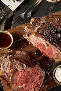 A perfectly cooked, juicy roast beef steak on a wooden cutting board with sides of dipping sauces and a glass of red wine.