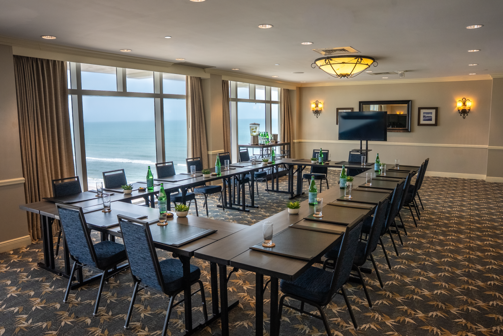 A conference room with tables, chairs, a large screen, and an ocean view through big windows.