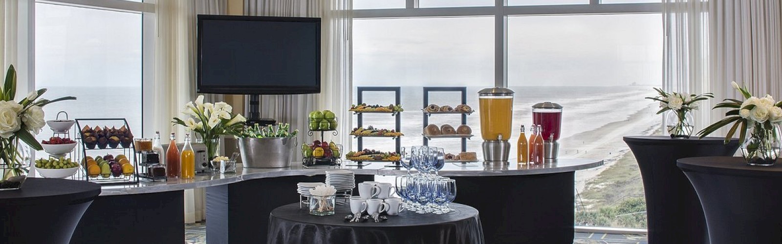 The image shows a well-arranged conference room with tables set for refreshments, including drinks, snacks, and floral decorations by large windows.