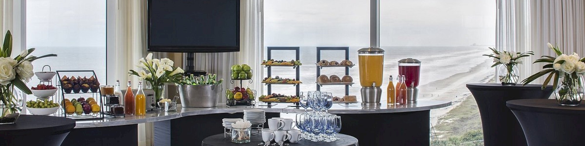 The image shows a well-arranged conference room with tables set for refreshments, including drinks, snacks, and floral decorations by large windows.