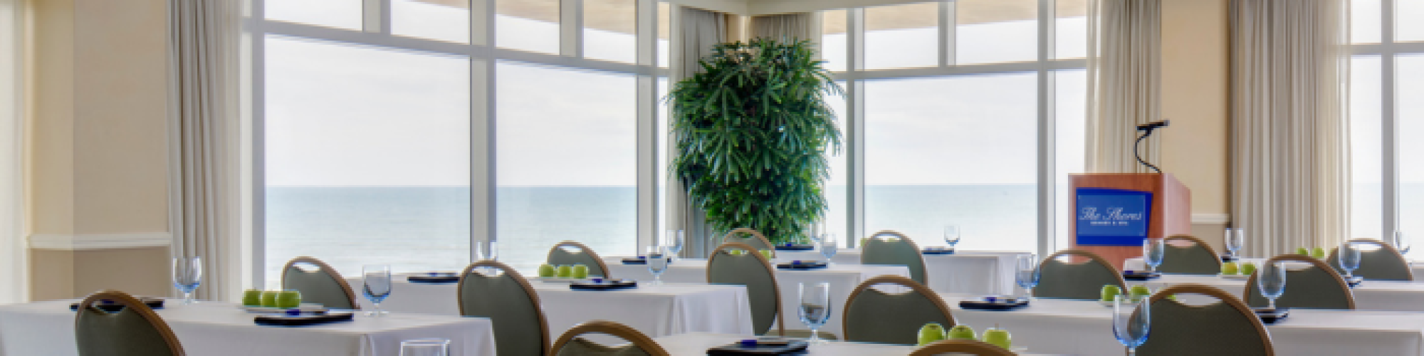 The image shows a conference room setup with tables, chairs, green apples, notepads, and glasses, overlooking a scenic ocean view through large windows.