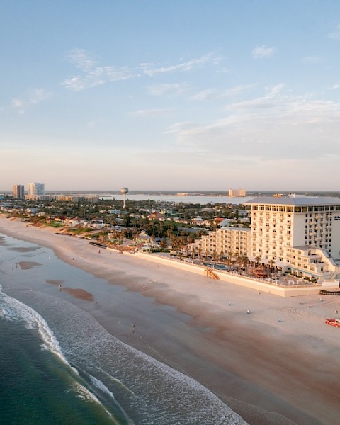 An aerial view of a beachside city with buildings, a sandy shore, and calm ocean waves during sunset.