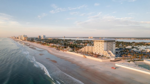 An aerial view of a beachside city with buildings, a sandy shore, and calm ocean waves during sunset.