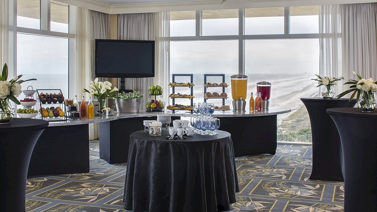 A conference room with a buffet setup including drinks and snacks, decorated with flowers and overlooking a coastal view through large windows.