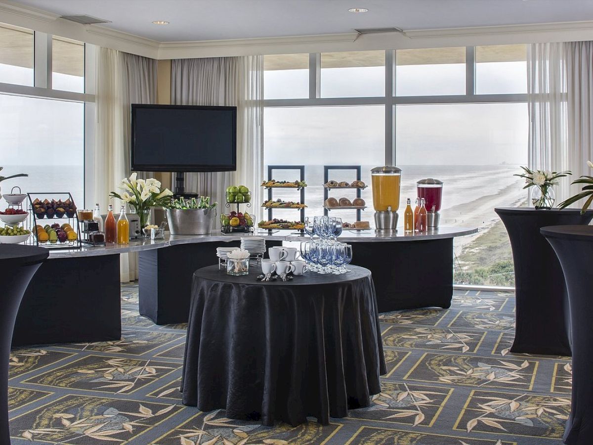 A conference room with a buffet setup including drinks and snacks, decorated with flowers and overlooking a coastal view through large windows.