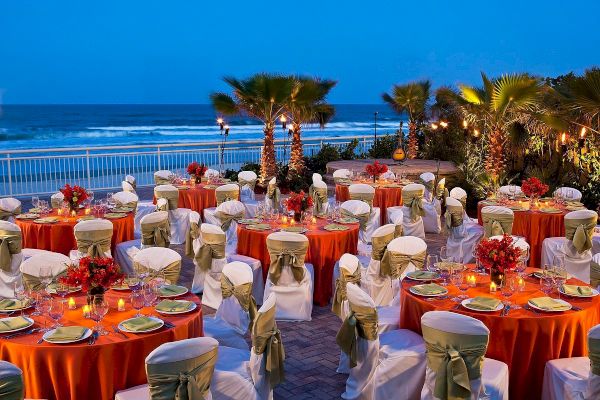 This image shows an outdoor event setup by the ocean, featuring round tables with vibrant orange tablecloths, adorned with floral centerpieces and elegant chairs.
