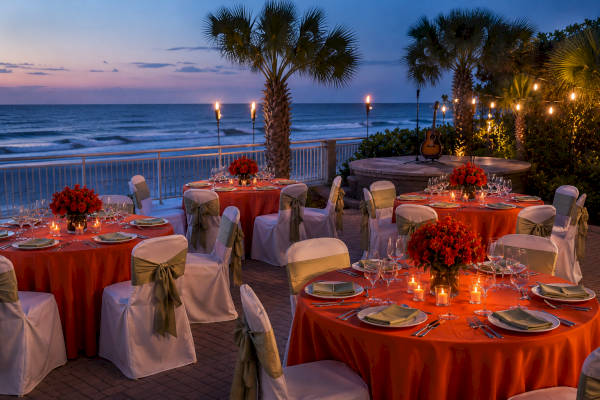 An outdoor beachside dinner setup with decorated tables, flowers, candles, palm trees, a guitar, and ocean views at dusk.