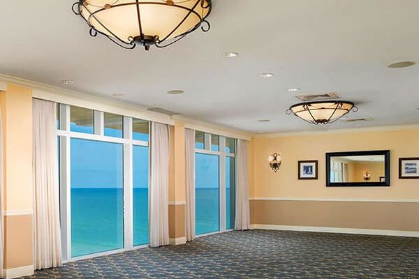 A spacious room with large windows offering an ocean view, elegant light fixtures, a mirror, and framed pictures on the walls, beige walls, and carpeted floor.
