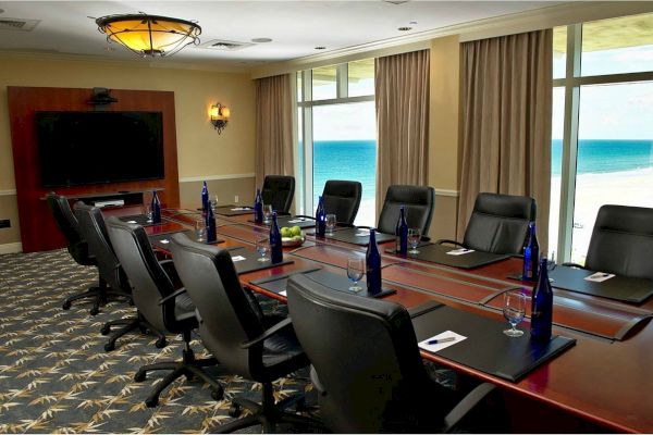 A conference room with a large table, empty chairs, water bottles, a bowl of green apples, a large TV, and a view of the ocean visible through windows.