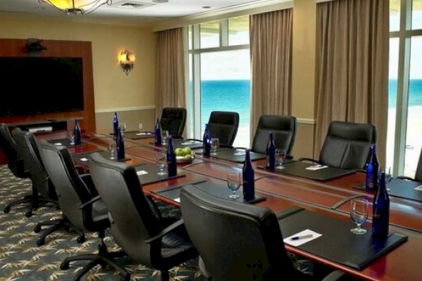 A conference room with a long table, black chairs, blue bottles, notepads, and a view of the ocean through large windows.