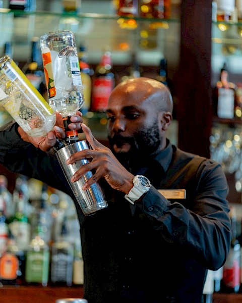A bartender pours a clear liquid into a shaker in a bar with shelves of colorful bottles behind him.