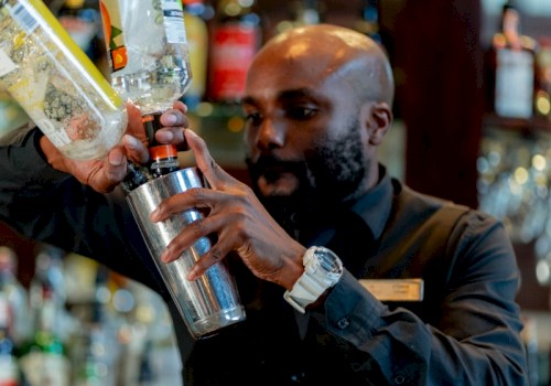 A bartender pours a clear liquid into a shaker in a bar with shelves of colorful bottles behind him.