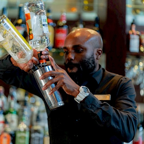 A bartender pours a clear liquid into a shaker in a bar with shelves of colorful bottles behind him.