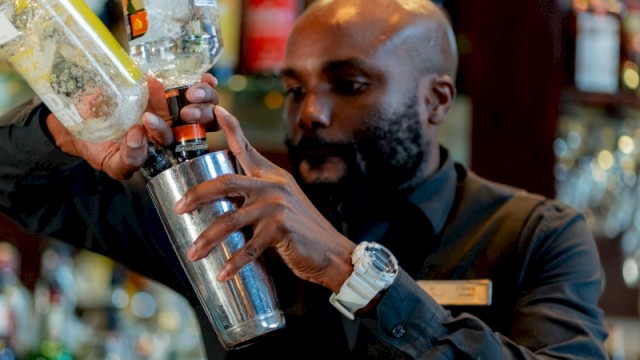 A bartender pours a clear liquid into a shaker in a bar with shelves of colorful bottles behind him.