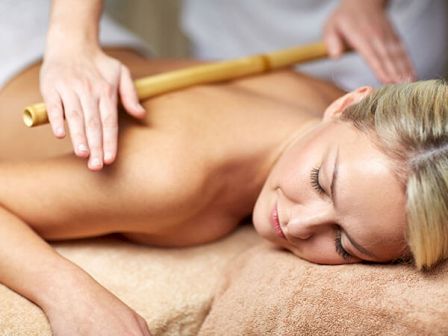 A person is lying on a massage table with a masseuse using a bamboo stick to perform a massage on their back.